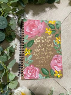 a spiral notebook with pink flowers and bible verse on the cover, surrounded by greenery