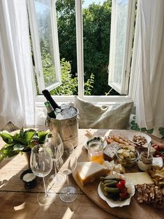 a wooden table topped with lots of food and wine next to two large open windows