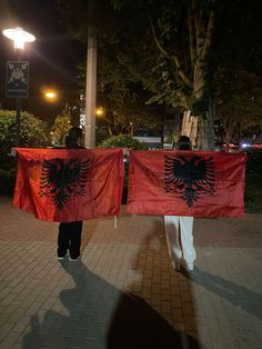 two people walking down the street holding red flags with black eagle on them and one person in white pants