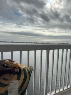 A hand holding a Starbucks apple croissant in its wrapper, with a scenic ocean view and cloudy sky in the background, seen from a white balcony railing.