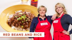 two women in red aprons standing next to a yellow plate with beans and rice