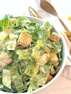 a salad with lettuce and croutons in a white bowl next to gold utensils