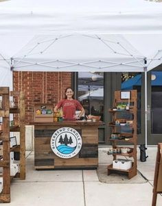 a woman standing behind a counter in front of a white tent with wooden shelves on it