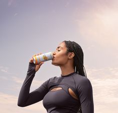 a woman drinking from a can while standing in front of a blue and white sky