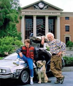 two men and a dog sitting in front of a car