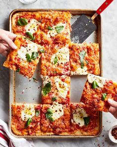 several square pieces of pizza with cheese and basil on top, being cut into squares