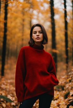 a woman standing in the middle of a forest with leaves on the ground and wearing a red sweater