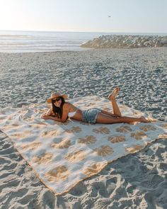 a woman laying on top of a sandy beach next to the ocean wearing a hat