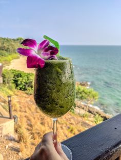 a person holding up a drink in front of the ocean with purple flowers on it