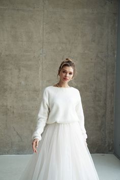 a woman standing in front of a concrete wall wearing a white dress with long sleeves