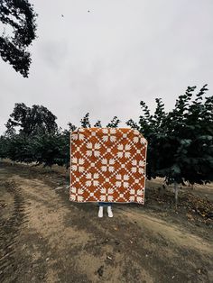 a person standing in the middle of an orchard holding up a large orange and white quilt