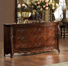 an ornate wooden dresser with flowers and vases on it's top, in front of a large mirror