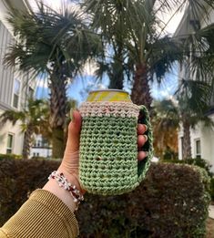 a hand holding a green crocheted coffee cup in front of a palm tree