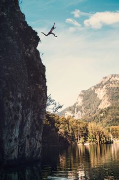 a person jumping off the edge of a cliff into a lake with mountains in the background