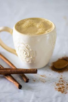 two cinnamon sticks next to a cup of coffee on a white tablecloth with cinnamons