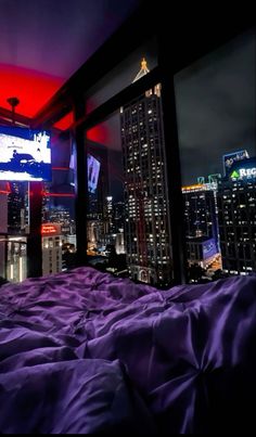 a bed with purple comforter in front of a city skyline at night, as seen from a bedroom window