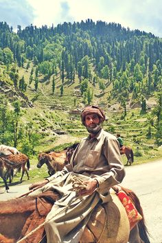 a man riding on the back of a brown horse down a road next to mountains