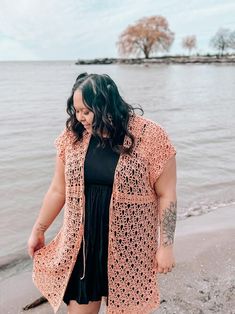 a woman standing on top of a beach next to the ocean wearing a pink crocheted cardigan