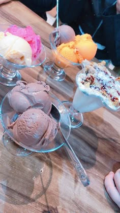 three different kinds of ice cream in glass dishes on a wooden table with spoons