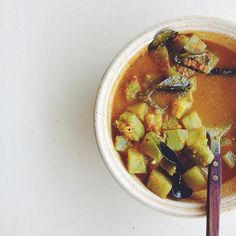 a bowl filled with soup and vegetables on top of a white table next to a purple spoon