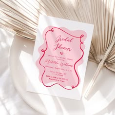 a pink and red wedding card sitting on top of a white plate next to a fan