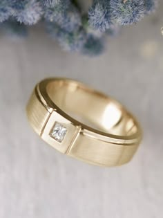 a yellow gold wedding ring with a princess cut diamond set in the center, sitting next to blue flowers