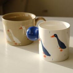 two coffee mugs sitting on top of a table next to each other with ducks painted on them