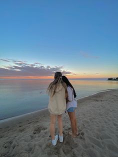 two girls are standing on the beach at sunset