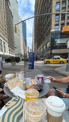 people sitting at an outdoor table with food and drinks in front of tall buildings on a city street