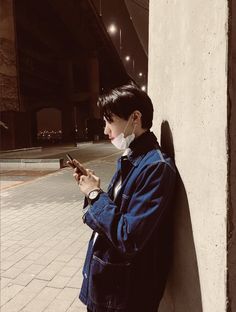 a woman leaning against a wall looking at her cell phone while wearing a face mask