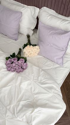 a bed with white sheets and purple pillows on it, next to a bouquet of flowers