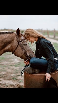 a woman sitting on top of a barrel petting a horse's head in a field