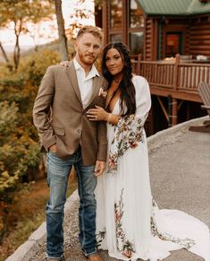 a man and woman standing next to each other in front of a log cabin at sunset