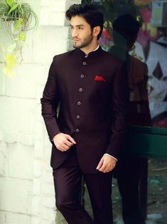 a man standing in front of a white brick wall wearing a maroon suit and tie