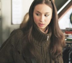 a woman with long brown hair wearing a sweater and looking at the camera while standing in front of a desk