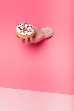 a hand holding a donut with chocolate sprinkles on it against a pink background