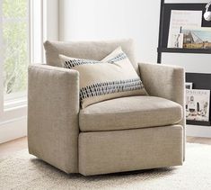 a chair with pillows on it in front of a window and framed pictures behind it