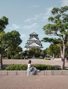 a woman sitting on the ground in front of trees and a building with a tower