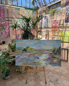 an easel is holding up a painting in a room filled with plants and paintings