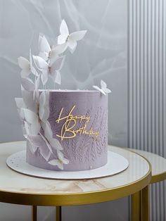 a purple birthday cake with white flowers and gold writing on it sitting on a table