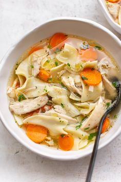 two bowls filled with chicken noodle soup on top of a white table next to a silver spoon