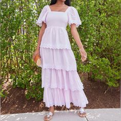 a woman wearing a white dress standing on a sidewalk in front of bushes and trees