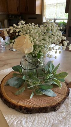 white flowers are in a glass vase on a wood slice that is sitting on a table