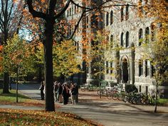 people walking down the sidewalk in front of an old building with autumn leaves on the ground