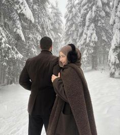 a man and woman standing in the snow next to some trees with their arms wrapped around each other