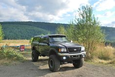 a large black truck parked on top of a dirt road next to a lush green forest