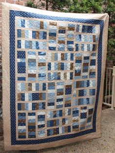 a large blue and tan quilt hanging on a brick wall next to a fence with trees in the background