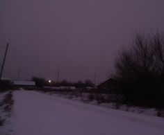 a snowy road with trees and buildings in the background at night, near an industrial area