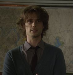 a man with long hair wearing a sweater and tie standing in front of a map