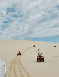 three people on four wheelers driving in the sand with cloudy sky behind them,